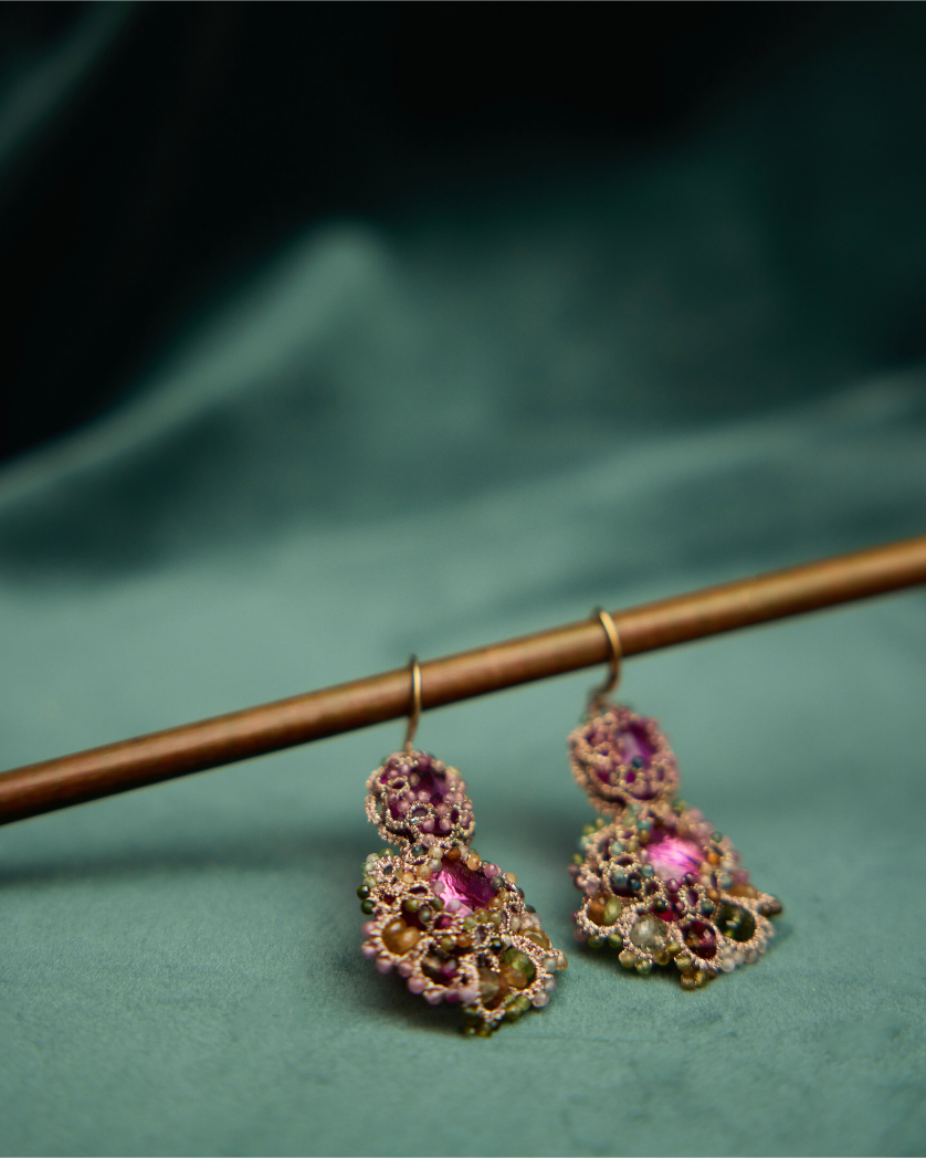 Rose silk earrings with intricate woven design, featuring pink and amber gemstones, displayed on a dark green fabric.