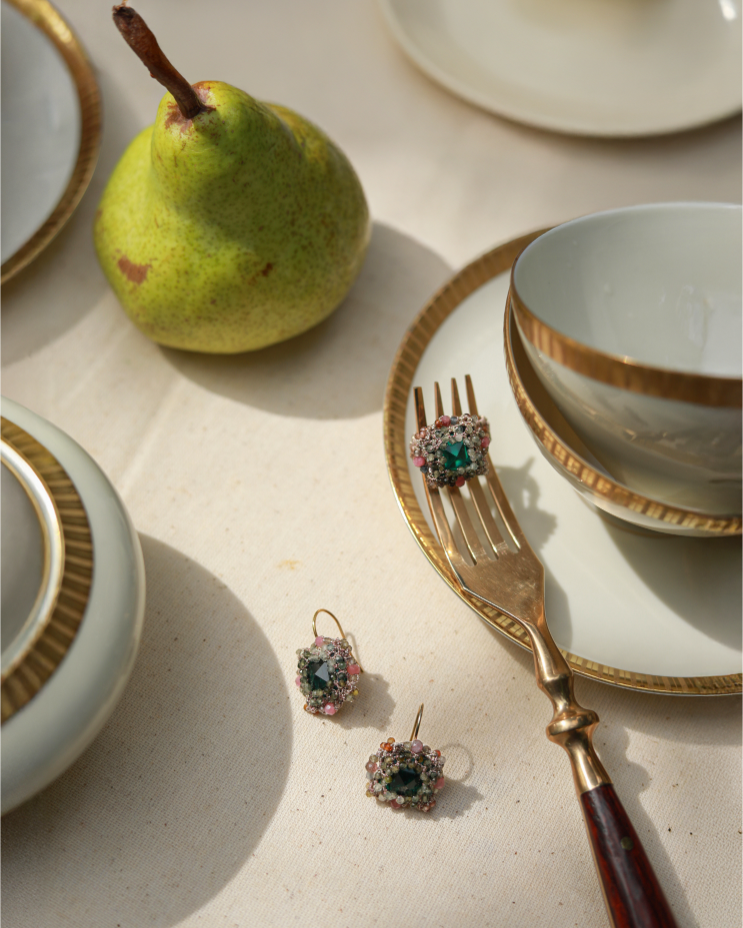 Silk ring with green crystal, pink tourmaline, and sapphire details on gold-rimmed tableware setting.