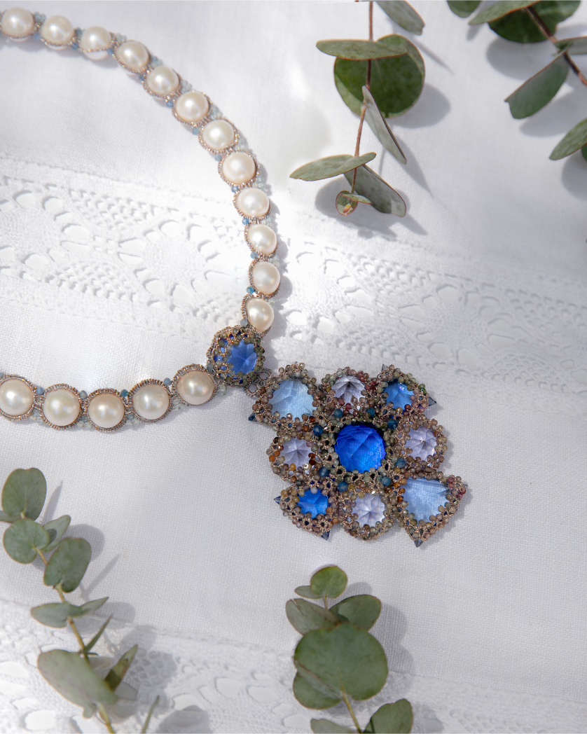 Pearl and blue gemstone floral pendant necklace on lace fabric with eucalyptus leaves.