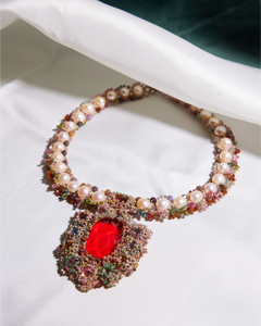 Silk necklace with pearls and multicolored beads, featuring a large red central stone on a white fabric background.