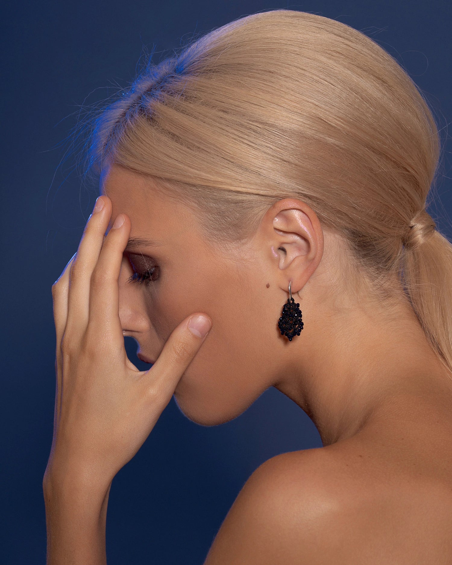 Black beaded DORA EARRINGS with silver hook, elegant side profile view against a rich blue background.