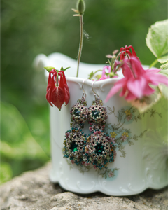 Ornate green earrings with intricate beadwork hanging on floral porcelain vase with flowers in background.
