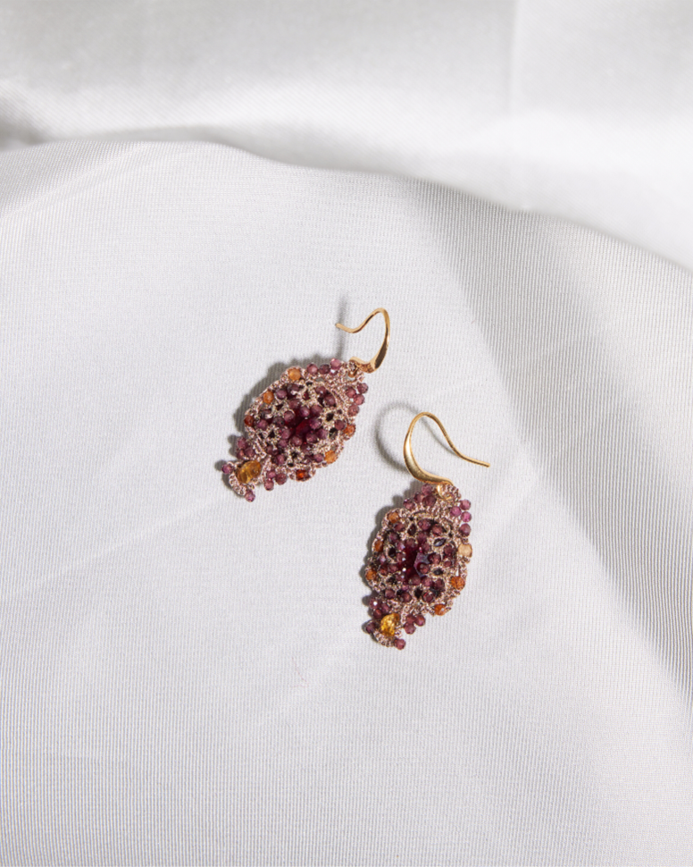 Gold hook earrings with intricate red and pink gemstone design on a textured white fabric.