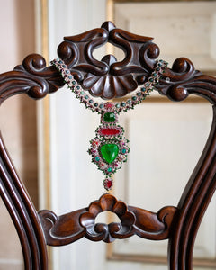 Elegant necklace with green crystal, red garnet, pearls, and intricate detailing, displayed on carved wooden chair.