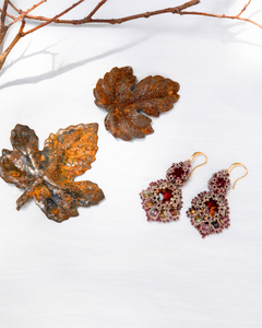 Red silk earrings with intricate beading and gold hooks on white fabric, paired with decorative metal leaves.