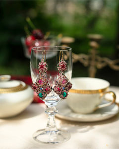 Ornate silk earrings with red and green gemstones displayed on a crystal glass, intricate texture detail.