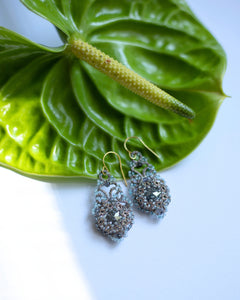 Blue Valentina earrings with intricate beading and gold hooks, side view on green leaf background.